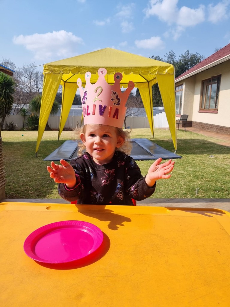 A joyful child wearing a paper crown, arms raised and smiling, outside in the sunshine at Brite Beginnings